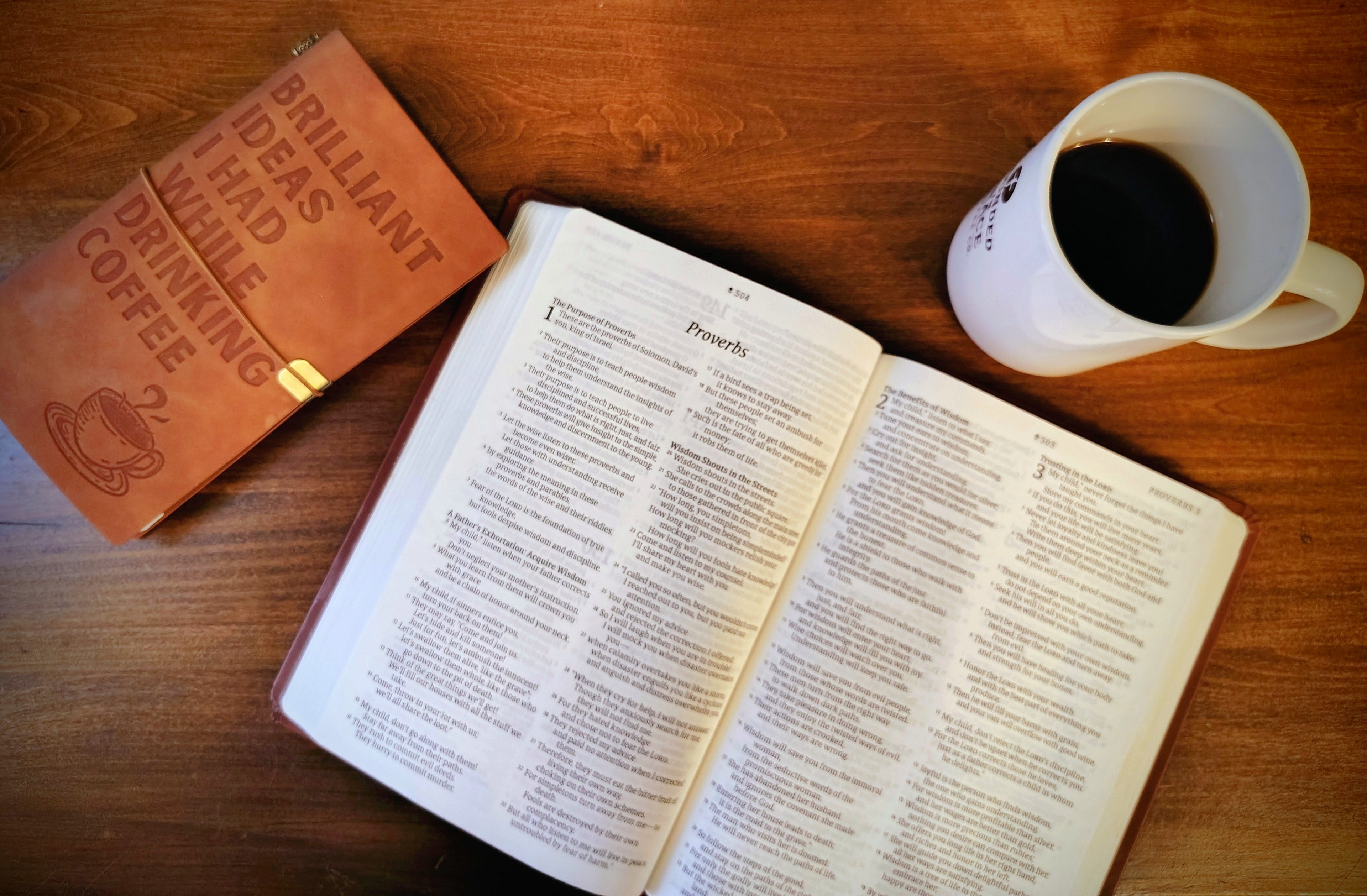 Open book on a wooden table with a coffee cup and leather notebook.